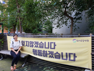 A young woman wearing a white shirt and blue jeans is sitting on a bench in front of a sign in Korean.