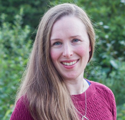A headshot of a person with long hair swept to the side, wearing a magenta sweater with a simply silver necklace.
