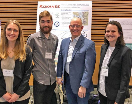 Four individuals wearing name badges around their necks and dressed in business attire, stand in front of a poster about their research.