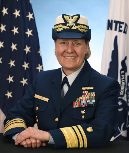 A woman in a US Coast Guard uniform poses in front of the US and Coast Guard flags.