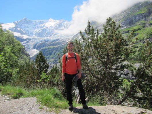 A man wearing athletic clothes is seen standing on a vista with a snow capped mountain peak in the background.
