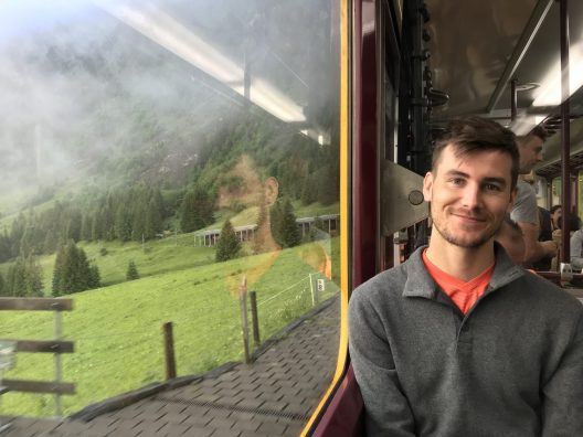 A man wearing a gray fleece with a red shirt underneathsits near the window inside a train. The scenery outside of the train is a foggy hillside with green grass fields in the foreground.