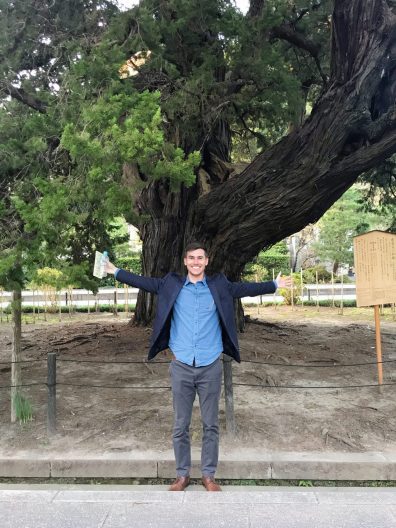 A man wearing a blue button up shirt under a navy sportcoat stands with arms outstretched wide in front of a large tree.