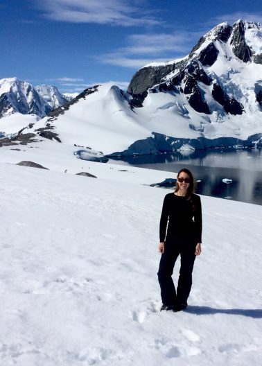 A woman is seen standing in the snow with dark gray, cold-looking water and a rocky, snowy mountain against a bright blue sky in the background.