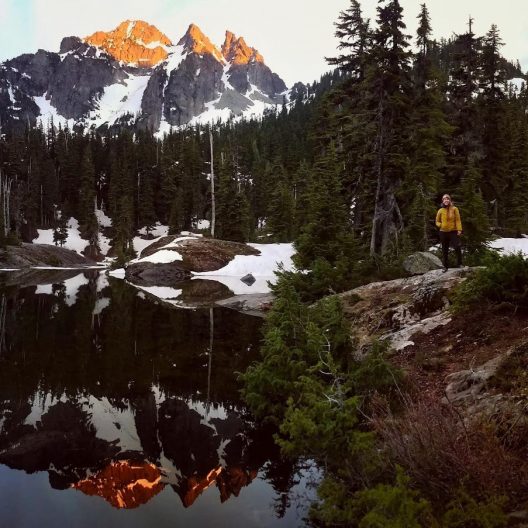 A woman wearing a yellow jacket is seen in the distance with a jagged, rocky mountain range rising out of the background. The mountain's reflection is seen in a flat waterbody in the foreground.