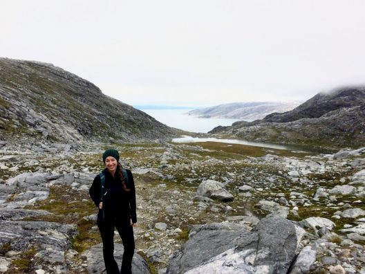 A woman stands on a rocky and mossy path wearing all black, a backpack, and a green knit hat. The ocean and rocky cliffs can be seen in the background