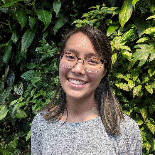 A headshot of Leslie Nguyen, wearing glasses and a gray top. She's standing in front of green foliage.