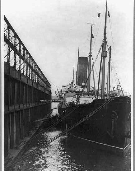  A steamship docked alongside a pier. Lines hold it in place, with two gangways leading to the upper and lower levels of the pier, respectively.