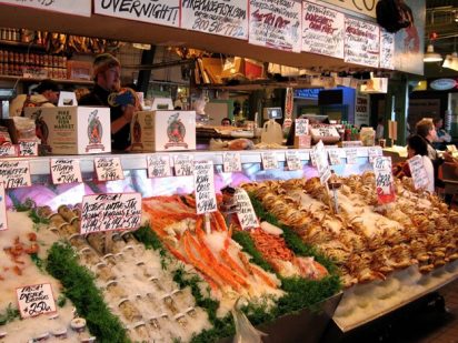 The storefront of a fish market, with products lined up in front of the vendors. The products include processed oysters in jars and various crabs all on ice. Written signs label the products and include their prices, as well as advertising other fish and promotions available to consumers.