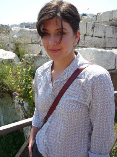 A woman wearing a light colored button up shirt with a cross-body bag stands in front of a partial wall of ruins in Turkey.
