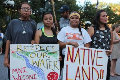four Indigenous youth stand in the foreground of the photo holding two signs, one reading "rezpect our water; mni wiconi" and the other reads "Native land!!"