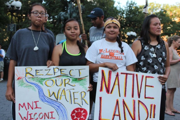 four Indigenous youth stand in the foreground of the photo holding two signs, one reading "rezpect our water; mni wiconi" and the other reads "Native land!!"