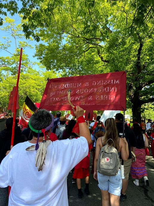 taken from behind, image shows a person burning sweetgrass to honor the MMIW movement; a red banner is held reading "missing and murdered Indigenous women, girls, and two spirit"