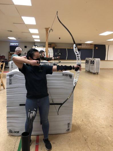 A woman dressed in jeans and a black t-shirt is standing, drawing back an arrow in her bow inside an indoor archery range.