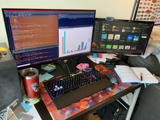 A workstation with multiple monitors, open notebook, pen, and coffee mug.