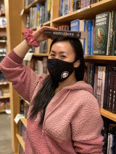 A young woman in a pink hoodie is balancing a hardcover book on her head in front of a row of bookshelves.