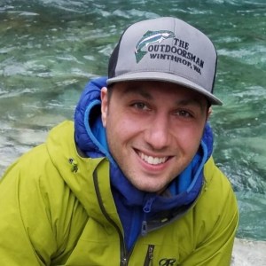 A headshot of Bobbak Talebi wearing a lime green jacket over a blue sweatshirt and wearing a gray baseball hat. He's standing in front of moving water.