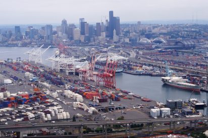 An aerial view of downtown Seattle's skyline. In the foreground, the mouth of a river is surround by cranes, boats, and shipping containers.