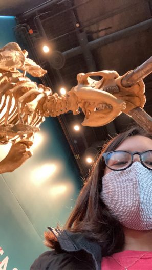 A masked Leah Huff takes a selfie looking upward toward the ceiling of the Burke Museum. Rising above her shoulder is the fossil of a wooly mammoth.