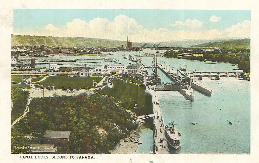 A colored photograph postcard of the Ballard Locks, showing the Locks on the right and Ballard on the left. One ship waits within the Locks to finish passing through, while another waits to be let in.