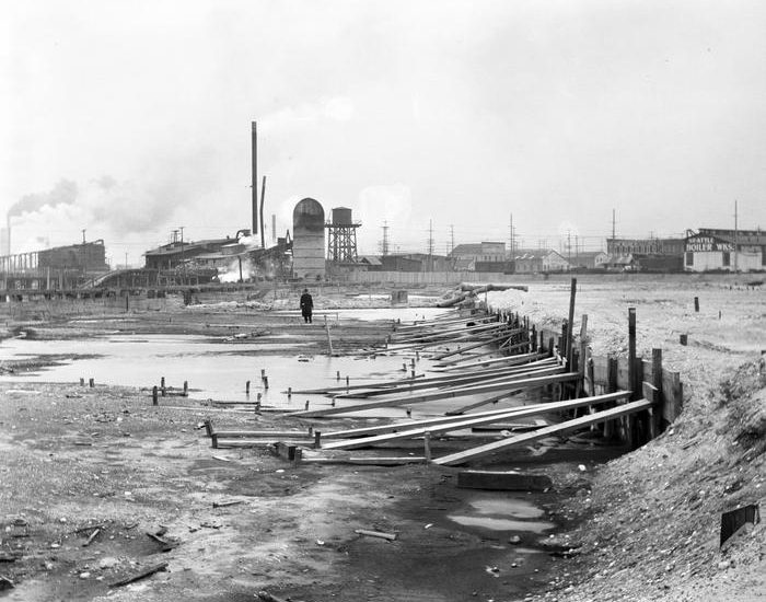 Wood planks hold up soil on the right side of the foreground, while in the center and left the ground remains partially flooded tidelands. In the background a person looks over the construction while behind him are warehouses, a silo, and a water tower.