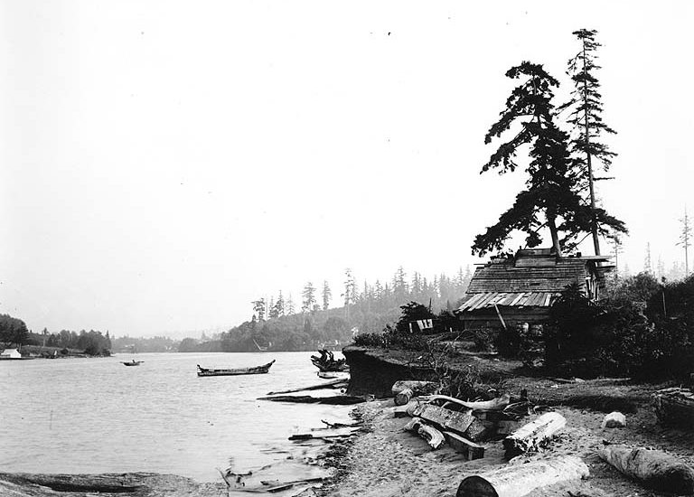 A cedar house on the banks of a river. A tree rises next to the house, and a canoe is tied nearby.