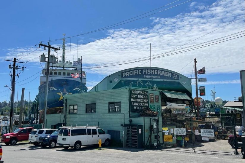 A green warehouse, seen from the front, with "Pacific Fishermen Inc. Shipyard" in white lettering. A fishing vessel is dry docked on the left side of the photo, awaiting repairs.