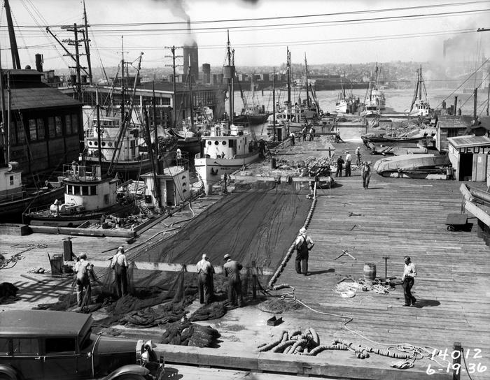 Vessels line one side of a dock, while on the dock people are working on a large fishing net.