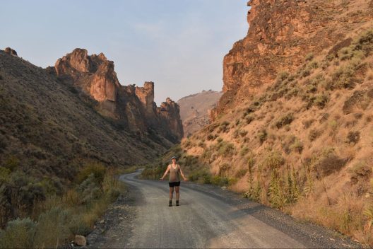 A woman with her arms out and a huge grin on her face stands in the middle of a gravel road that traverses an impressive rocky canyon lined with sagebrush.