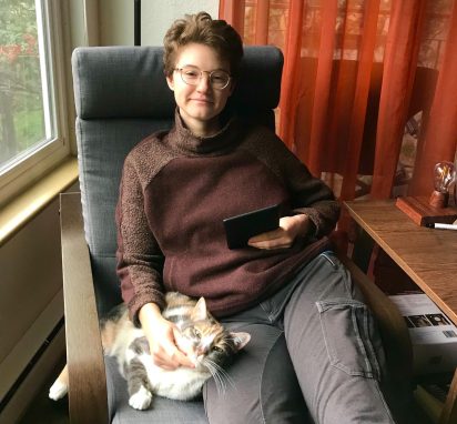 Author, Kat, is sitting by a window with their cat and holds a Kindle.