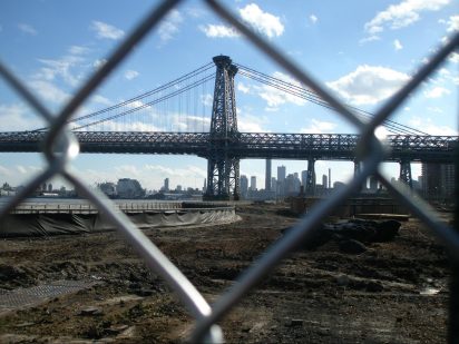 A view of the Williamsburg Bridge through a chain-link fence. Below the bridge is a dirt covered construction site where the East River Park once was.