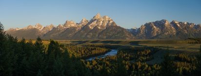 Under a blue sky at sunset, majestic mountain peaks tower above a vibrant pine forest, a sprawling grassy field, and the curving Snake River.