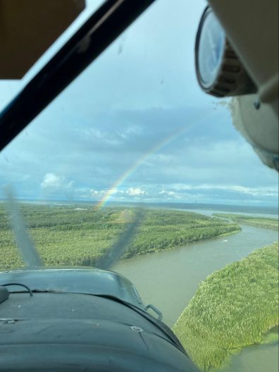 Through the front window of a small airplane, a river stretches below into the distance amongst forest. Just ahead, a rainbow arcs over a small runway.