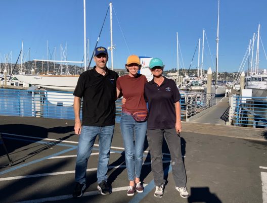 three people, each members of the team for this project, stand near a dock with fishing vessels behind them on a beautifully sunny day.