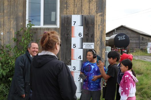 Three students stand with two adults in front of a building that has a flood guage attached to it. The students are recording the adults talking.