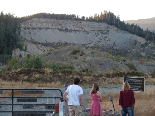 Three people stand in front of the memorial at the Oso Mudslide site. It's dusk, and before them lies a huge heap of sediment that has fallen from the hill behind it.