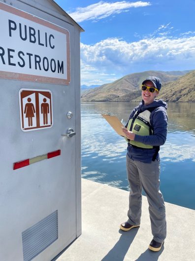 A person wearing a life jacket and cap smiles while holding a clipboard near a public restroom by a calm lake, with hills and a blue sky in the background.