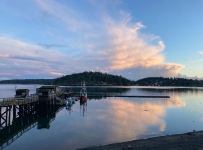 A view of a dock extending into the glassy waters of the Salish Sea with forested land across the channel. Wispy sunlit clouds dance in the sky.