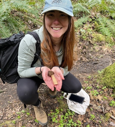 Kneeling on a forest floor, Jazzmin excitedly and proudly showing off a verpa mushroom.