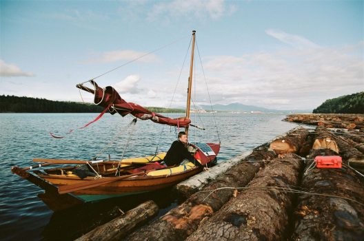 The small double ended dory is floating alongside gigantic log booms that stretch to the horizon. Smoke rises for a factory in the far distance.