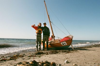 The author and his friend, Pax stand besides their disheveled boat pulled up on a beach. Both have their thumbs down and drysuits on.