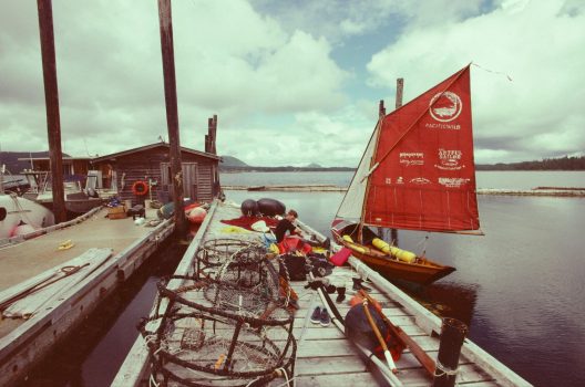 Two docks parallel to each other. The dory with its full sail out drying is tied to a listing dock. The Pacific Wild logo of an intertwined bear and salmon is fully visible on its sail. Amidst fishing equipment on the dock Pax packs up a mess of gear for the ride home.