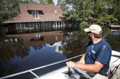 In the background dark flood waters from Hurricane Florence rise up the trunks of trees and to a few feet below the roofline of a house. A shirtless man stands with his hands on his hips. In the foreground, a man with a baseball cap drives a small motorboat through the floodwaters.