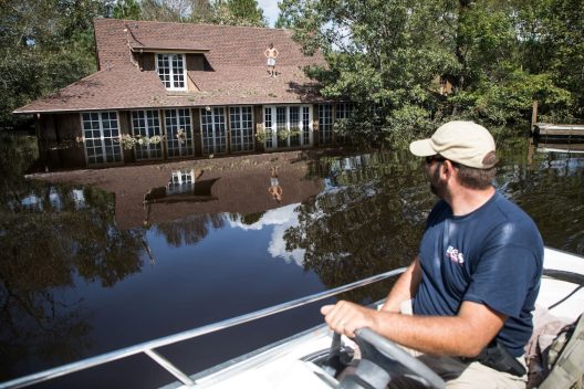 In the background dark flood waters from Hurricane Florence rise up the trunks of trees and to a few feet below the roofline of a house. A shirtless man stands with his hands on his hips. In the foreground, a man with a baseball cap drives a small motorboat through the floodwaters.