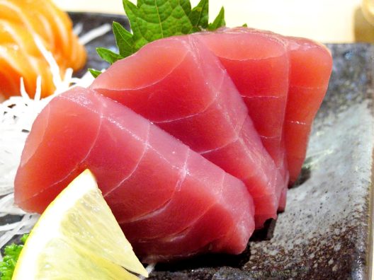4 pieces of bright red maguro sashimi on a plate with a slice of lemon and a shiso leaf for decoration.