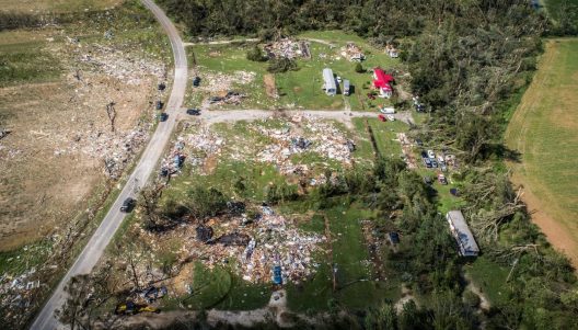 An aerial view of a road, an agricultural field, some forested land and the remains of a trailer park that has been demolished by a tornado. Remnants of the trailers and the items inside are strew haphazardly towards the top left of the image. Some vehicles are flipped over. Three trailer appear unscathed and some cars are parked on the road and amongst the wreckage.