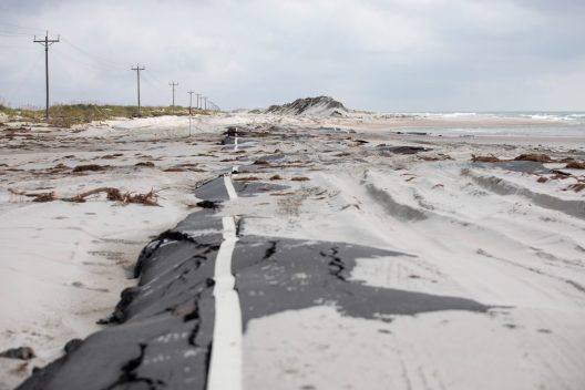 Powerlines, a road, sand dunes and beach extend into the distance. The line of tall dunes is broken and shows the ocean, the beach, and where a storm surge broke through the dunes. The road is buckled and covered in sand. There are tire tracks from cars or trucks in the sand that covers the road.