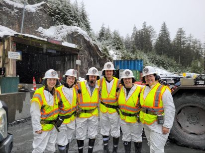 Five capstone students and their capstone client stand at the entryway into Greens Creek mine, at the base of a large, snow-capped mountain.