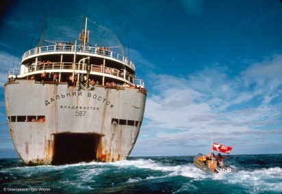 On the open ocean, a large industrial Soviet whaling boat is approached by a small zodiac operated by Greenpeace during a protest. The greenpeace boat is seen waving a flag.