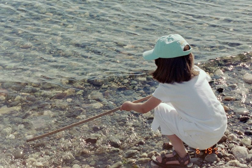 A young girl squatting down on a rocky shoreline using a stick to poke at the water's edge.